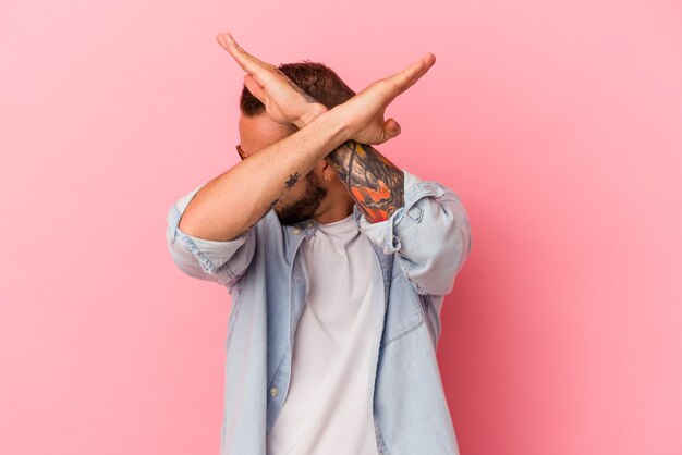Giovane uomo caucasico con tatuaggi isolati su sfondo rosa mantenendo due braccia incrociate, concetto di negazione.