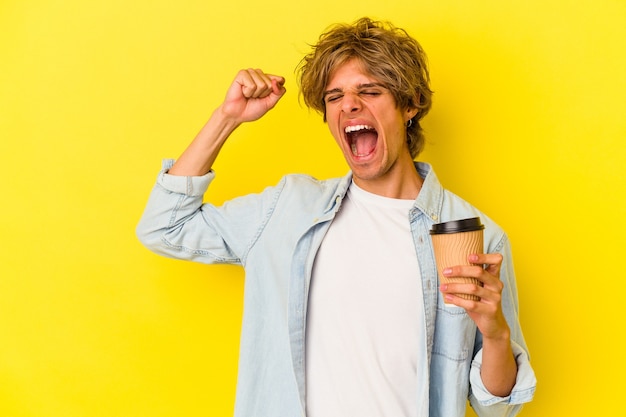 Giovane uomo caucasico con trucco che tiene un caffè da asporto isolato su sfondo giallo alzando il pugno dopo una vittoria, concetto vincitore.