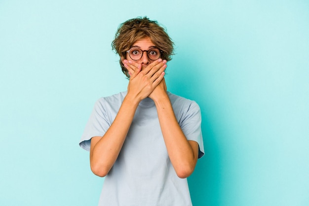 Giovane uomo caucasico con trucco isolato su sfondo blu scioccato che copre la bocca con le mani.