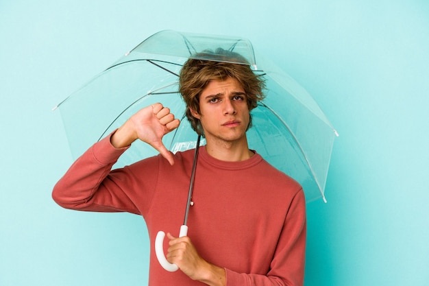 Giovane uomo caucasico con trucco tenendo l'ombrello isolato su sfondo blu che mostra un gesto di antipatia, pollice in giù. Concetto di disaccordo.