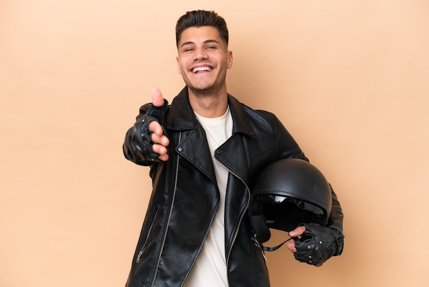 Giovane uomo caucasico con un casco da motociclista isolato su sfondo beige che stringe la mano per chiudere un buon affare