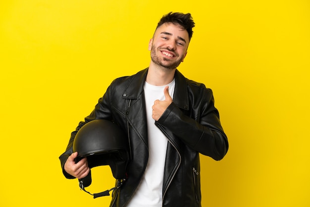 Giovane uomo caucasico con un casco da motociclista isolato su sfondo giallo che dà un gesto di pollice in alto