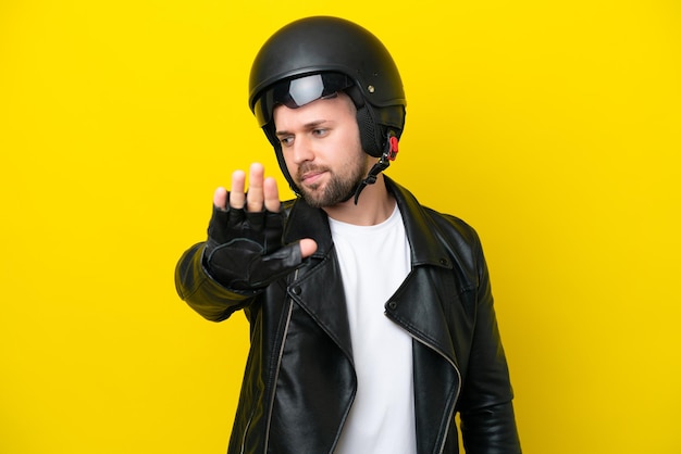 Giovane uomo caucasico con un casco da motociclista isolato su sfondo giallo che fa un gesto di arresto e deluso