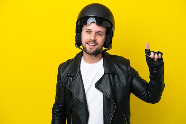 Giovane uomo caucasico con un casco da motociclista isolato su sfondo giallo che mostra e solleva un dito in segno del meglio