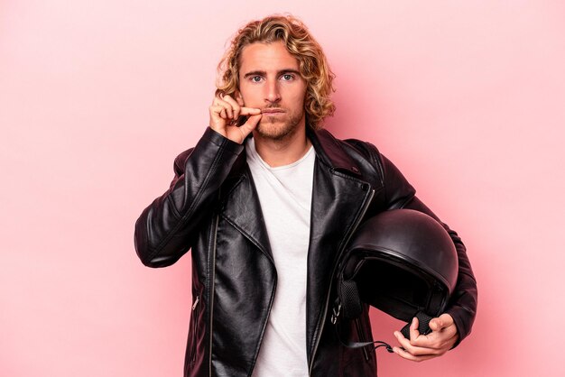 Giovane uomo caucasico con un casco da motociclista isolato su sfondo rosa con le dita sulle labbra che mantengono un segreto.