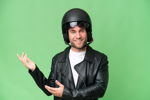 Giovane uomo caucasico con un casco da motociclista isolato su sfondo verde croma che estende le mani di lato per invitare a venire