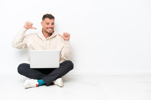 Giovane uomo caucasico con un computer portatile seduto sul pavimento isolato su sfondo bianco orgoglioso e soddisfatto di sé