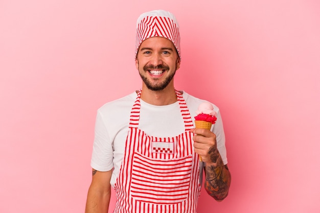 Giovane uomo caucasico del creatore del ghiaccio con i tatuaggi che tengono il gelato isolato su fondo rosa felice, sorridente e allegro.