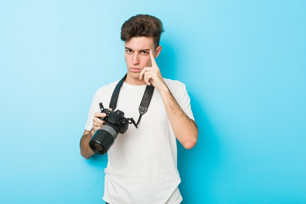 Giovane uomo caucasico del fotografo che tiene una macchina fotografica che tocca il suo tempio con il dito