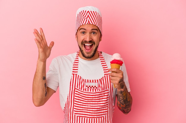 Giovane uomo caucasico del produttore di ghiaccio con tatuaggi che tengono gelato isolato su sfondo rosa ricevendo una piacevole sorpresa, eccitato e alzando le mani.