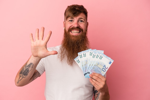 Giovane uomo caucasico dello zenzero con la barba lunga che tiene le banconote isolate su fondo rosa sorridente allegro che mostra il numero cinque con le dita.