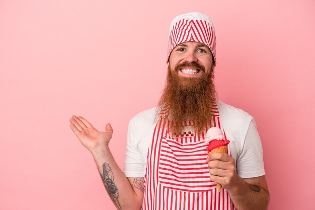 Giovane uomo caucasico dello zenzero con la barba lunga che tiene un gelato isolato su sfondo rosa che mostra uno spazio di copia su un palmo e tiene un'altra mano sulla vita.