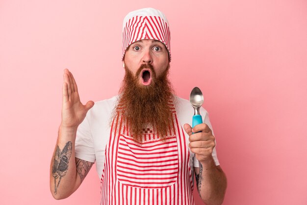 Giovane uomo caucasico dello zenzero con la barba lunga che tiene una paletta isolata su fondo rosa sorpreso e scioccato.