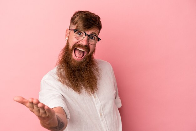 Giovane uomo caucasico dello zenzero con la barba lunga isolata su fondo rosa che allunga la mano alla macchina fotografica nel gesto di saluto.