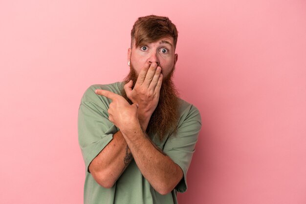 Giovane uomo caucasico dello zenzero con la barba lunga isolata su fondo rosa che indica il lato