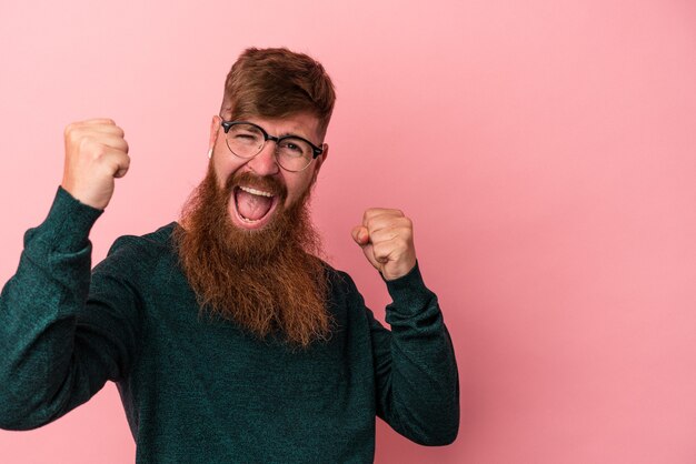 Giovane uomo caucasico dello zenzero con la barba lunga isolato su sfondo rosa tifo spensierato ed eccitato. Concetto di vittoria.