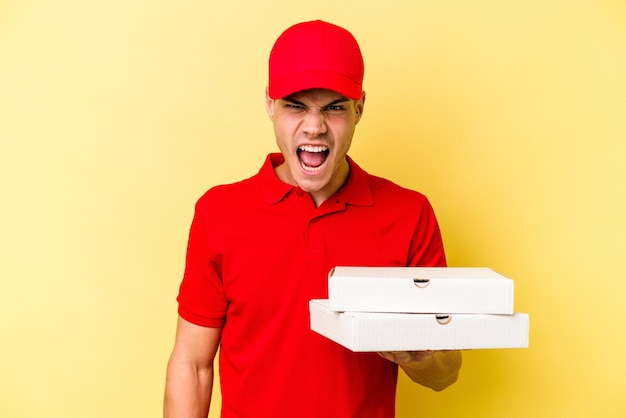 Giovane uomo caucasico di consegna che tiene pizze isolate su sfondo giallo che urla molto arrabbiato e aggressivo.