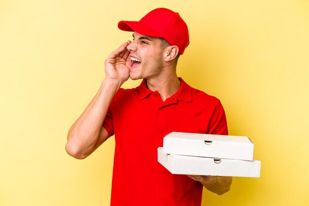 Giovane uomo caucasico di consegna che tiene pizze isolate su sfondo giallo gridando e tenendo il palmo vicino alla bocca aperta