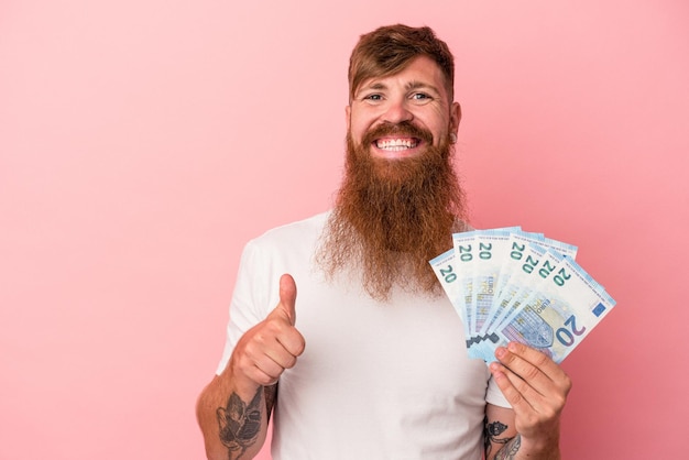 Giovane uomo caucasico di zenzero con barba lunga che tiene banconote isolate su sfondo rosa sorridente e alzando il pollice