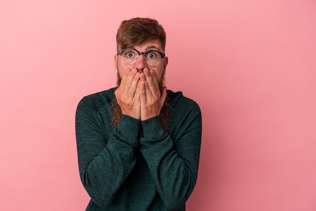 Giovane uomo caucasico di zenzero con barba lunga isolato su sfondo rosa scioccato, che copre la bocca con le mani, ansioso di scoprire qualcosa di nuovo.