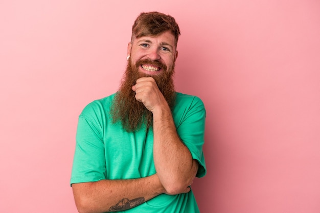 Giovane uomo caucasico di zenzero con barba lunga isolato su sfondo rosa sorridente felice e fiducioso, toccando il mento con la mano.