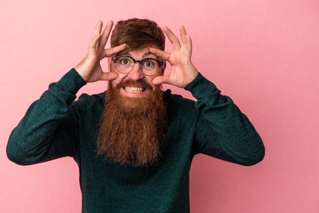 Giovane uomo caucasico di zenzero con barba lunga isolato su sfondo rosa tenendo gli occhi aperti per trovare un'opportunità di successo.