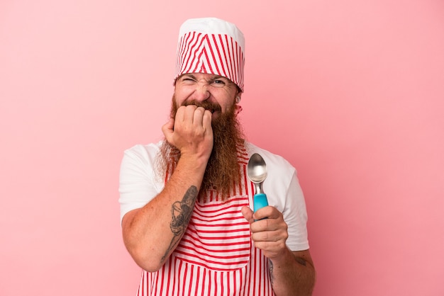 Giovane uomo caucasico di zenzero con la barba lunga che tiene in mano un mestolo isolato su sfondo rosa che si morde le unghie, nervoso e molto ansioso.