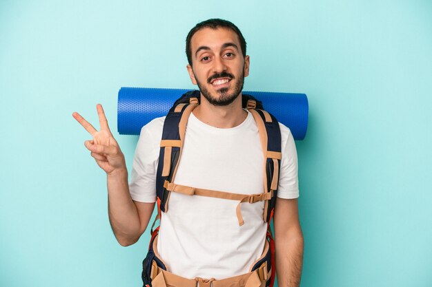 Giovane uomo caucasico escursionista isolato su sfondo blu gioioso e spensierato che mostra un simbolo di pace con le dita.