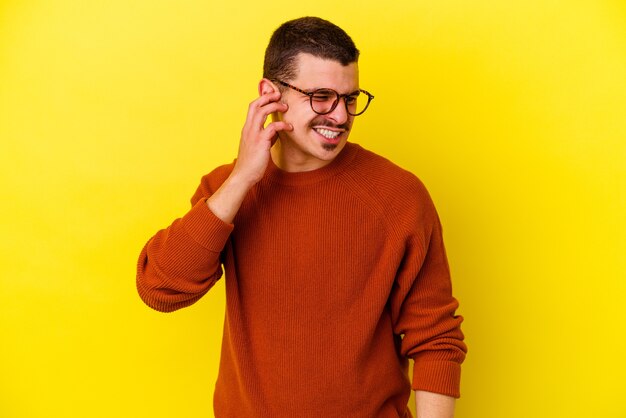 Giovane uomo caucasico freddo isolato sulle orecchie del rivestimento murale giallo con le mani.