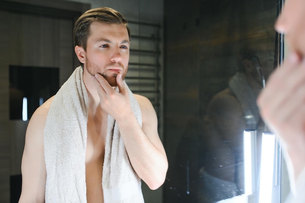 Giovane uomo caucasico guardarsi allo specchio fare la routine di cura della pelle del viso dopo la doccia nel bagno di casa maschio millenario in asciugamano dopo il bagno applicare lozione o crema per il viso sulla pelle dopo la rasatura concetto di igiene