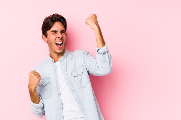 Giovane uomo caucasico in posa in un pugno di sollevamento isolato rosa dopo una vittoria, concetto di vincitore.