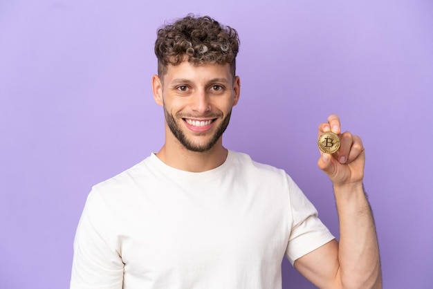 Giovane uomo caucasico in possesso di un Bitcoin isolato su sfondo viola in posa con le braccia all'anca e sorridente