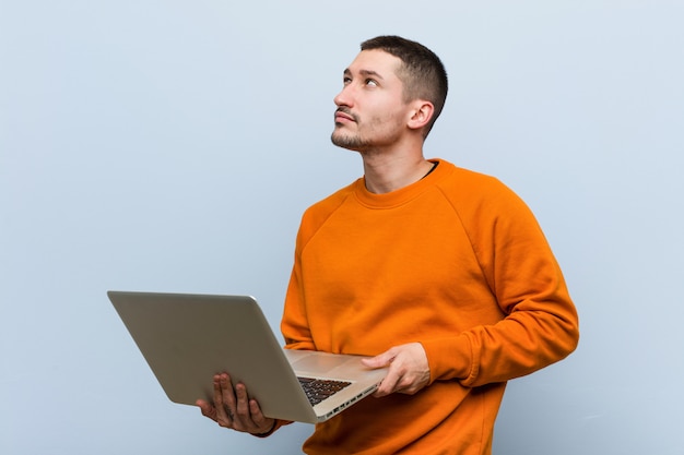 Giovane uomo caucasico in possesso di un computer portatile sorridente fiducioso con le braccia incrociate.