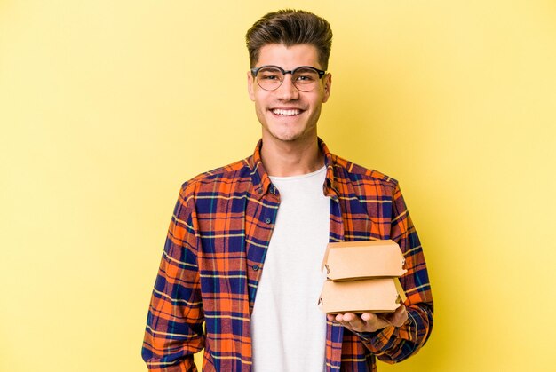 Giovane uomo caucasico in possesso di un hamburger isolato su sfondo giallo felice, sorridente e allegro.