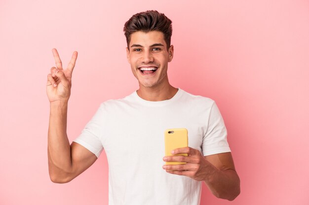 Giovane uomo caucasico in possesso di un telefono cellulare isolato su sfondo rosa gioioso e spensierato che mostra un simbolo di pace con le dita.