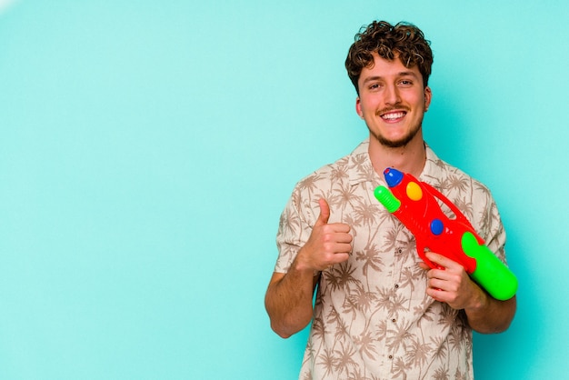 Giovane uomo caucasico in possesso di una pistola ad acqua isolata su sfondo blu sorridente e alzando il pollice