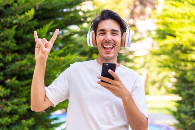 Giovane uomo caucasico in un parco che ascolta musica con un cellulare che fa gesto rock