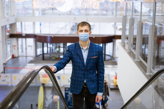 Giovane uomo caucasico in una giacca e una mascherina medica si arrampica sulla scala mobile in un centro commerciale