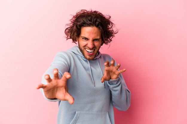 Giovane uomo caucasico isolato su bakcground rosa che mostra artigli che imitano un gatto, gesto aggressivo.