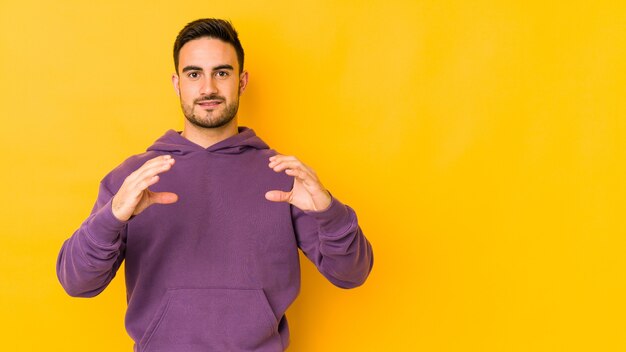 Giovane uomo caucasico isolato su giallo tenendo qualcosa con le palme