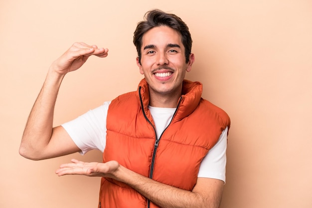 Giovane uomo caucasico isolato su sfondo beige che tiene qualcosa di piccolo con gli indici sorridenti e fiduciosi