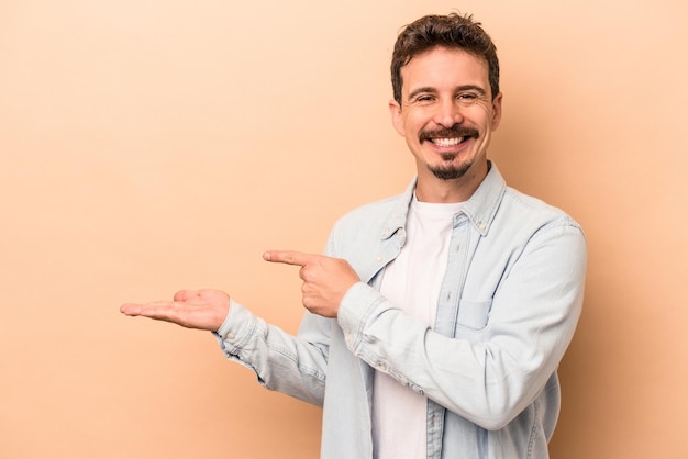 Giovane uomo caucasico isolato su sfondo beige eccitato in possesso di uno spazio di copia sul palmo.