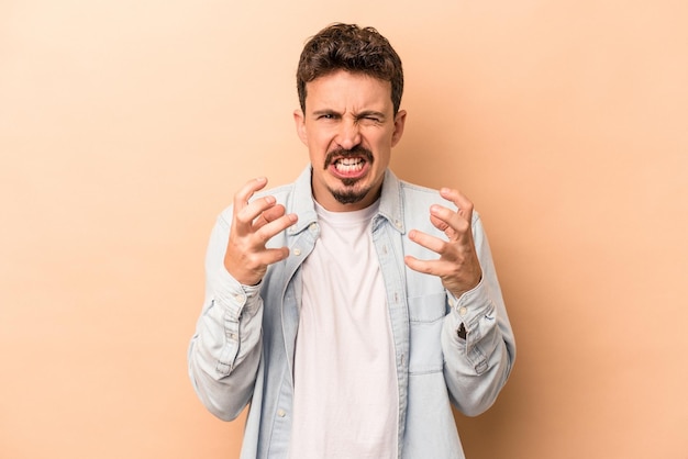 Giovane uomo caucasico isolato su sfondo beige sconvolto urlando con le mani tese.