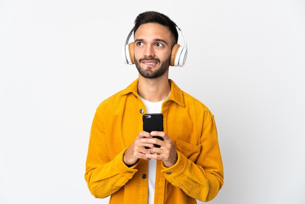 Giovane uomo caucasico isolato su sfondo bianco ascoltando musica con un cellulare e pensando