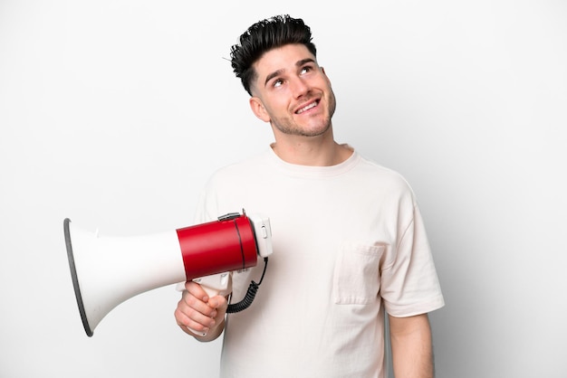 Giovane uomo caucasico isolato su sfondo bianco in possesso di un megafono e alzando lo sguardo mentre sorride