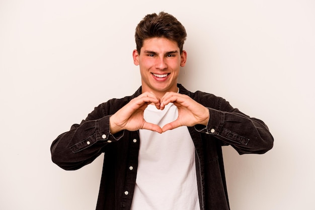 Giovane uomo caucasico isolato su sfondo bianco sorridente e mostrando una forma di cuore con le mani