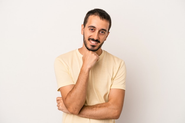 Giovane uomo caucasico isolato su sfondo bianco sorridente felice e fiducioso, toccando il mento con la mano.