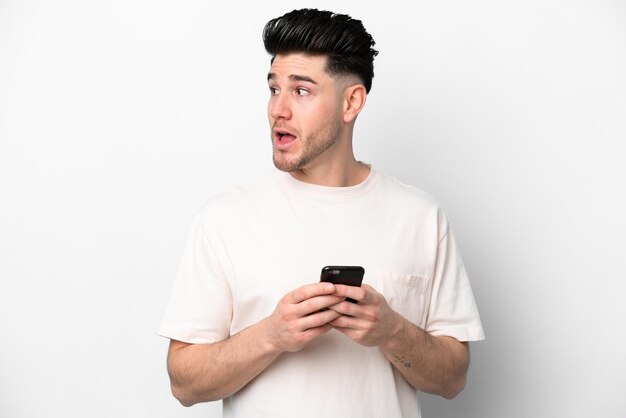 Giovane uomo caucasico isolato su sfondo bianco utilizzando il telefono cellulare e alzando lo sguardo