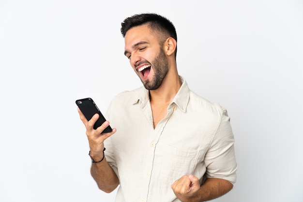 Giovane uomo caucasico isolato su sfondo bianco utilizzando il telefono cellulare e facendo gesto di vittoria