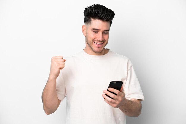 Giovane uomo caucasico isolato su sfondo bianco utilizzando il telefono cellulare e facendo gesto di vittoria
